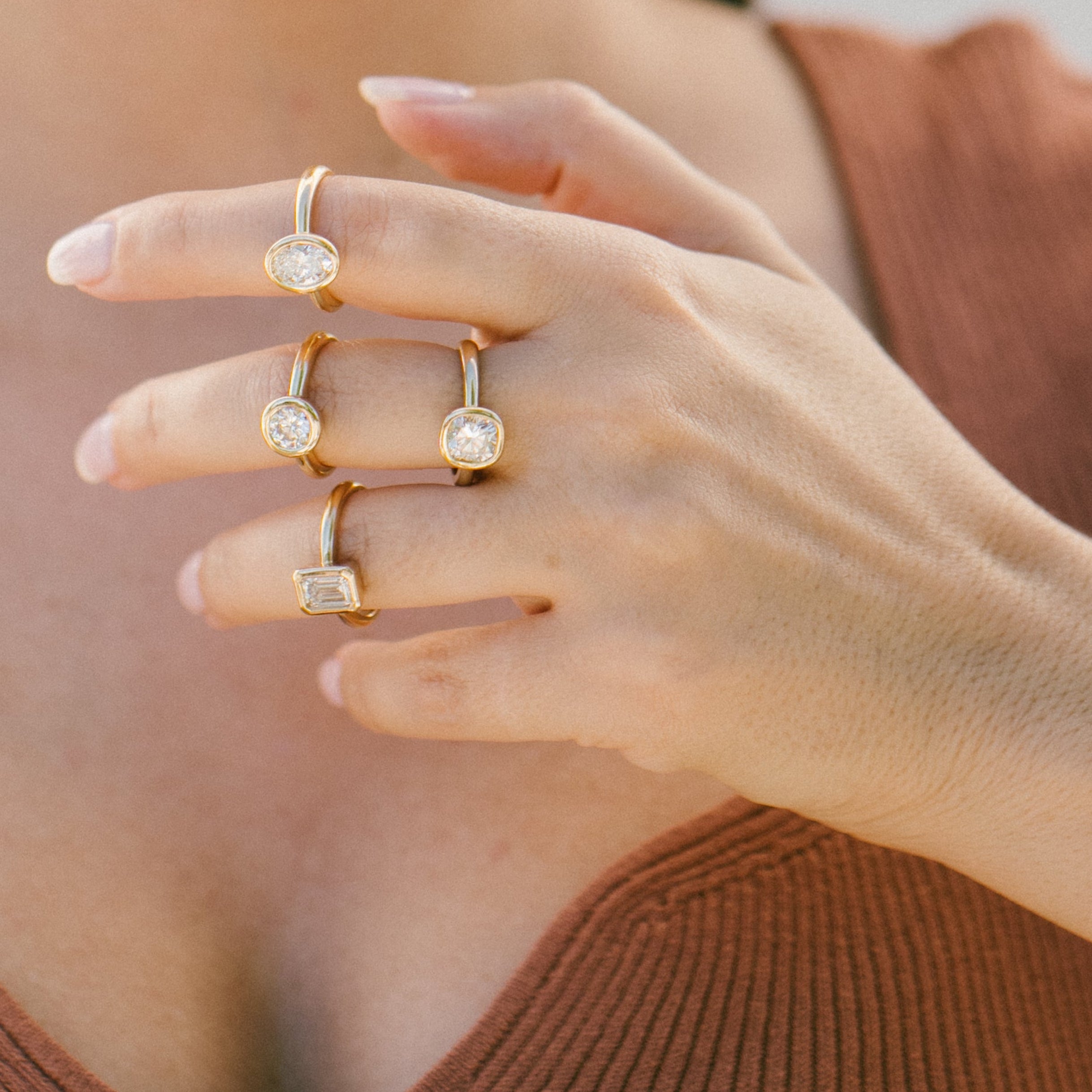 A close-up lifestyle shot of a person wearing four gold rings with clear gemstones on their fingers, wearing a terracotta-colored ribbed knit top.