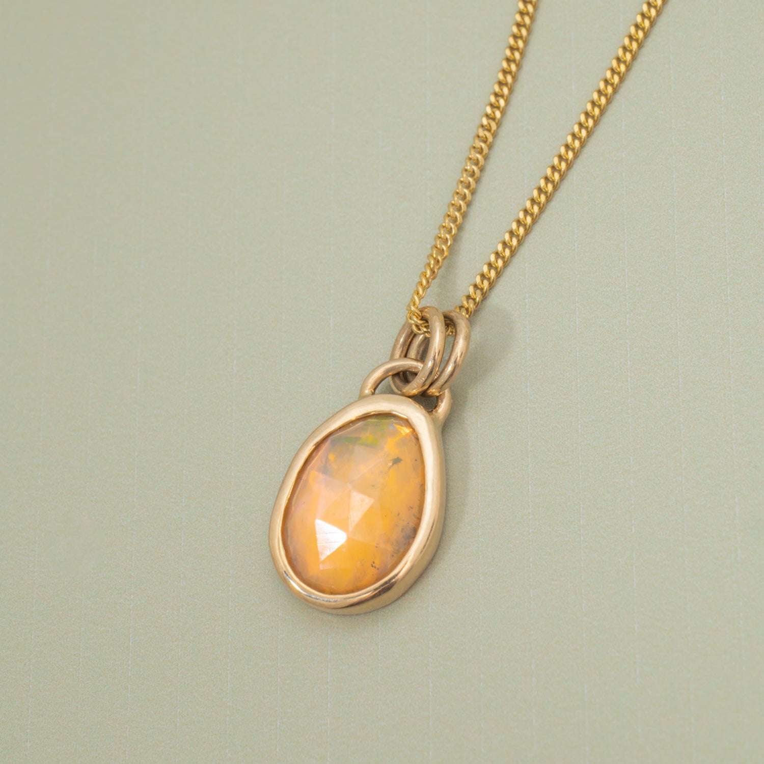 A gold chain necklace with a teardrop-shaped faceted opal pendant in a gold bezel setting, displayed on a textured light sage green background.