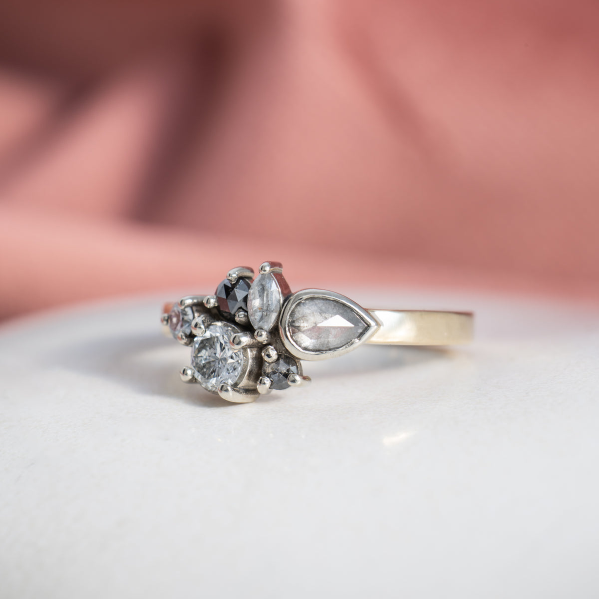 Susan cluster ring with white and grey stones on a yellow gold band, on white ceramic with pink backdrop