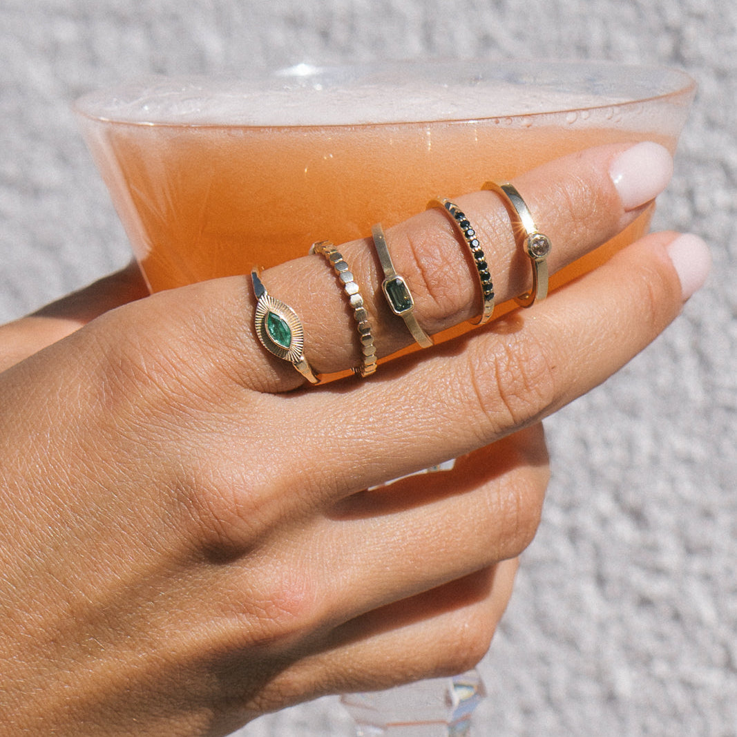 A close-up of a hand holding a glass of orange-colored drink, wearing five stacked gold rings with green and black accents.