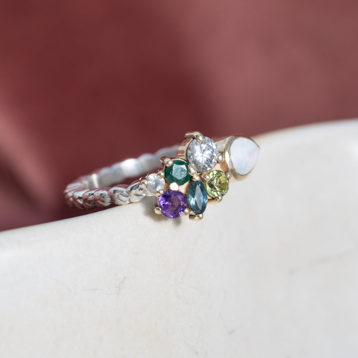 Cathy mixed metal cluster ring with colourful gemstones and an opal on white ceramic, burgundy backdrop
