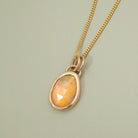 A gold chain necklace with a teardrop-shaped faceted opal pendant in a gold bezel setting, displayed on a textured light sage green background.